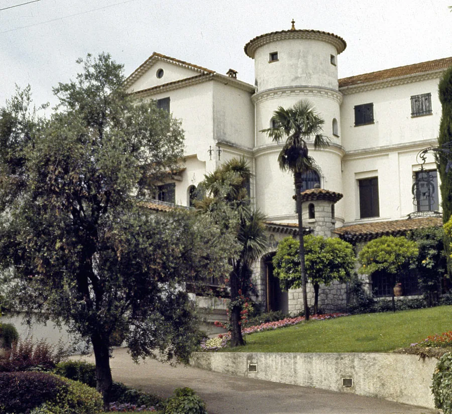 A white villa with a round corner tower and tiled roofs behind an olive tree, palms and a clipped lawn, photographed in Grasse.
